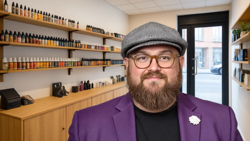 Bearded man in a flat cap and purple blazer standing in a modern vape shop with e-liquid bottles on wooden shelves